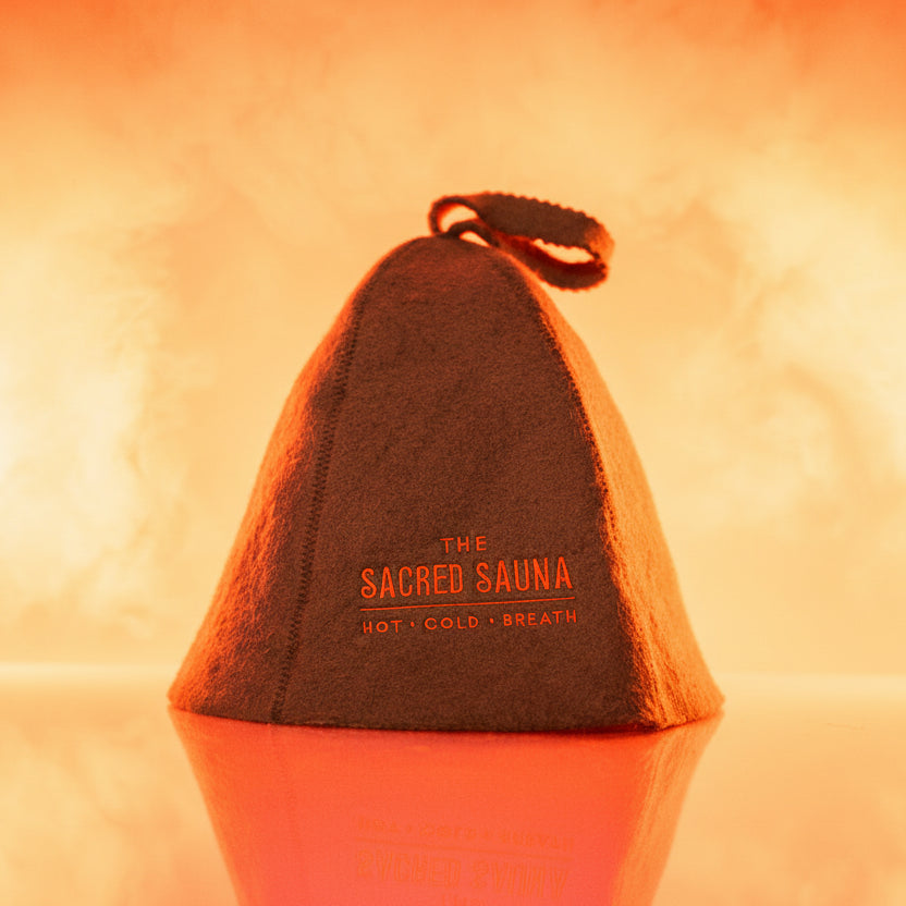 En grå bastumössa med logotypen 'The Sacred Sauna' i orange text. Mössan står på en reflekterande yta med en mjuk orange bakgrund, vilket skapar en varm och inbjudande atmosfär.