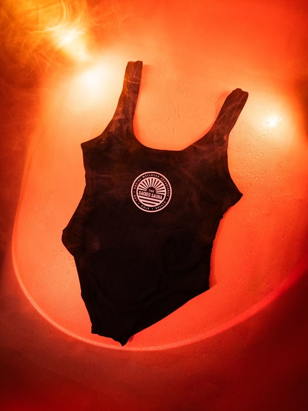 Svart baddräkt med The Sacred Sauna-logotyp på framsidan, hängande mot en fuktig yta med röd bakgrund och rök. Baddräkten är designad för bastubad och avkoppling.